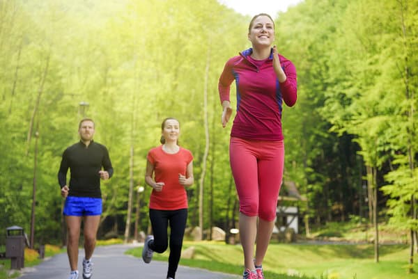 Three people running on a paved path surrounded by greenery. The woman in the foreground wears a red top and pink pants. Behind her, a woman in a red top and black pants, and a man in a black top and blue shorts are also running. Trees fill the background.