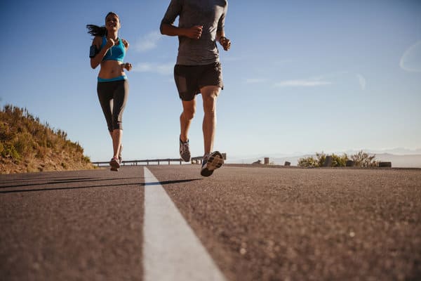 Two individuals are running outdoors on an open road. The person on the left is wearing a sports bra and leggings, while the person on the right is wearing a long-sleeved shirt and shorts. The sunny, clear sky and distant mountain range provide a scenic backdrop.