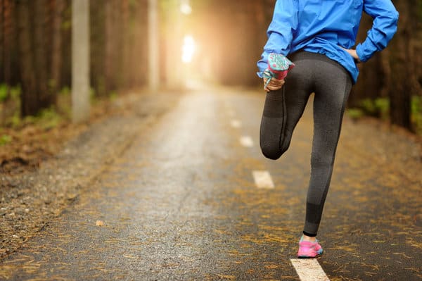 A person wearing a blue jacket, black leggings, and pink running shoes stretches by pulling one leg behind them on a forest path. Sunlight filters through trees, illuminating the scene with a warm glow.