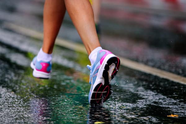 Close-up of an athlete's legs running on a wet road, wearing white socks and colorful running shoes in pink, blue, and white. The road reflects the light, indicating it has recently rained.