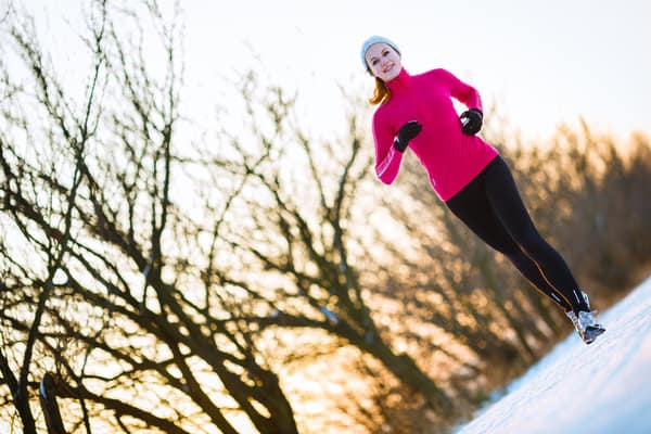 A person in a pink jacket and black leggings is jogging on a snow-covered path beside leafless trees. They are wearing a gray beanie and black gloves, and the sky is a gradient of warm colors suggesting sunrise or sunset.