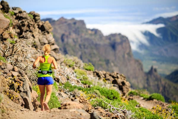 A person with a green backpack and blue shorts runs along a rocky trail surrounded by mountainous terrain and lush vegetation. The background features a panoramic view of mist-covered valleys and rugged peaks under a clear blue sky.