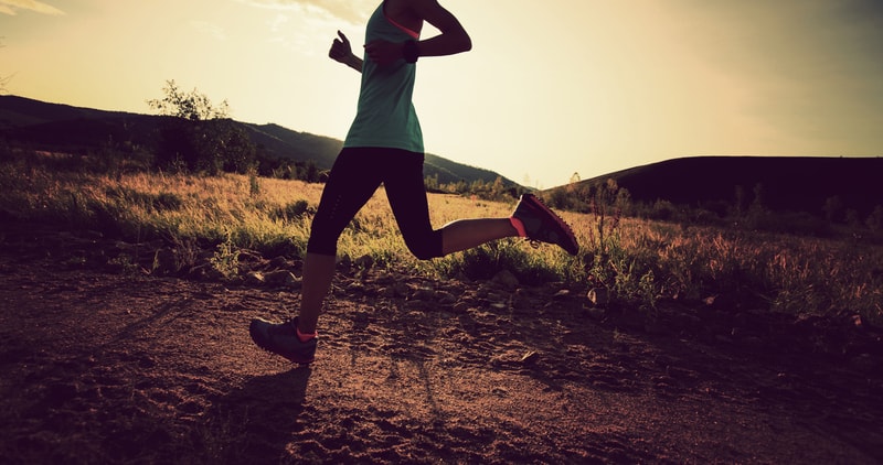 A person wearing athletic clothing and running shoes is jogging on a dirt path with a scenic backdrop of rolling hills and a setting sun. The silhouette of their legs and shoes is highlighted against the warm, golden light of the evening.