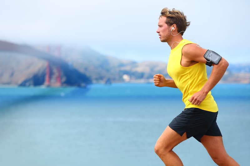 A man in a yellow tank top and black shorts jogs along a waterfront, wearing earphones and an armband. A large red bridge and mountains can be seen in the background under a cloudy sky.