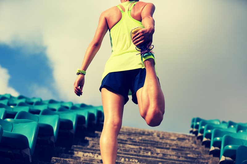 A person wearing a bright green tank top and black shorts is stretching their leg while standing on stadium steps. The person is grasping their foot behind them, preparing for a workout. The teal seats of the stadium and the cloudy sky are visible in the background.