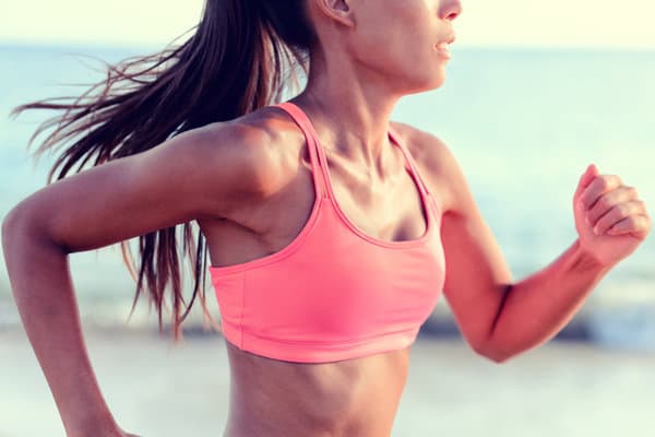 A person in a pink sports bra and ponytail jogging on a beach. The ocean and sky are visible in the background. Their face is partially out of frame, focusing on the upper body. The scene suggests a healthy and active lifestyle.