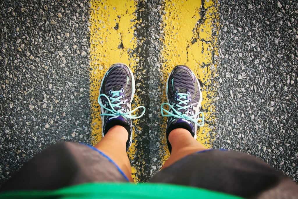 A person wearing blue and black running shoes with white laces stands on a textured asphalt road, positioned between two yellow lines. The individual is wearing black shorts and a green shirt, with their legs visible from above.