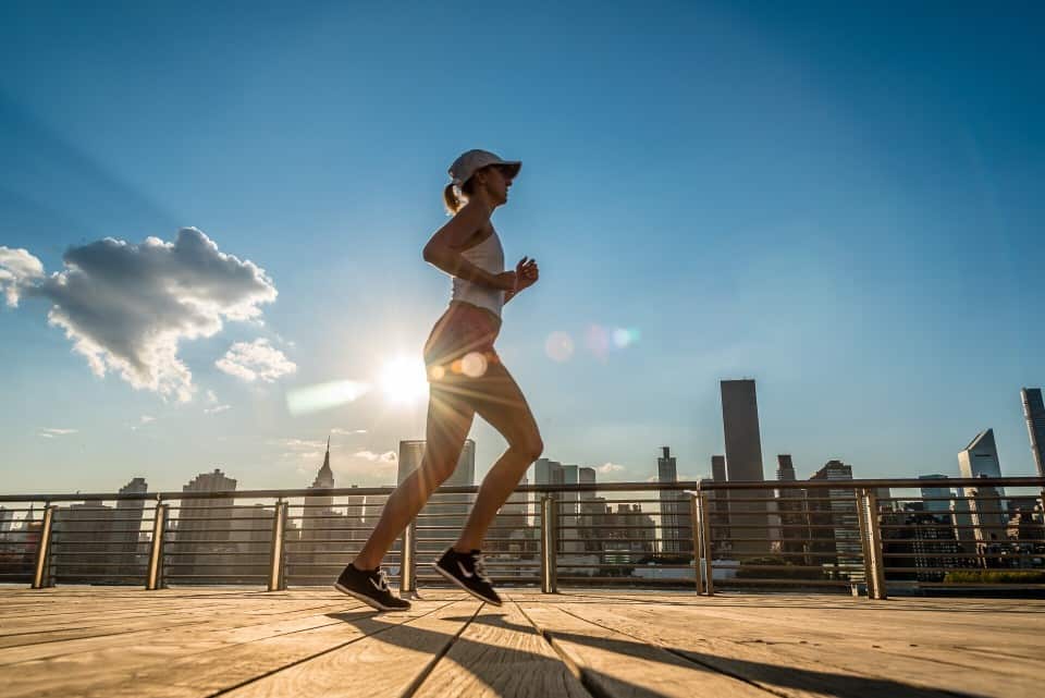 A person runs along a wooden boardwalk during sunset, with a city skyline and partly cloudy sky in the background. The runner is wearing a white cap, tank top, and sneakers, and the sun creates a bright flare behind them.