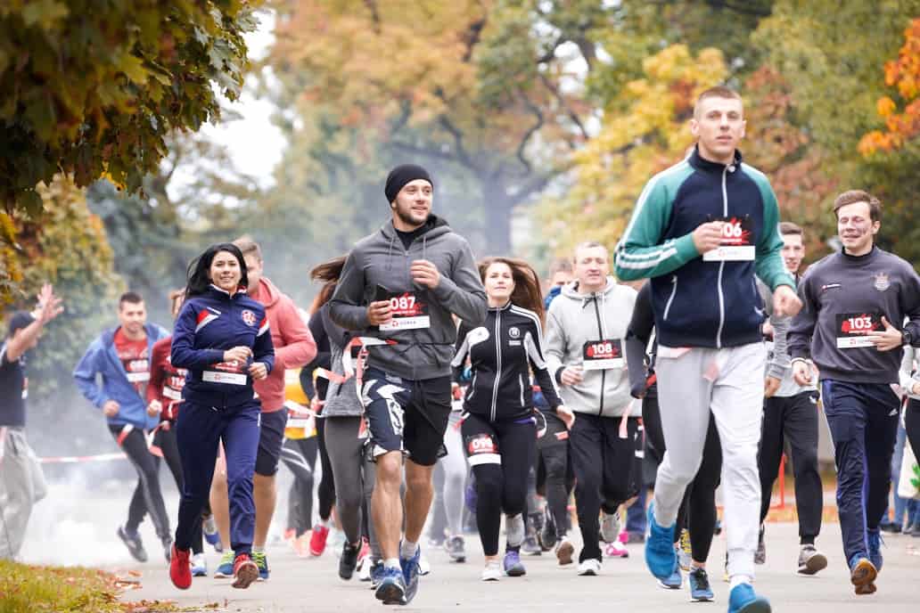 A group of people of varying ages and genders wearing athletic clothing are running in a park during a race. The background shows trees with autumn foliage. Most participants are focused on the race, and they have race numbers pinned to their attire.