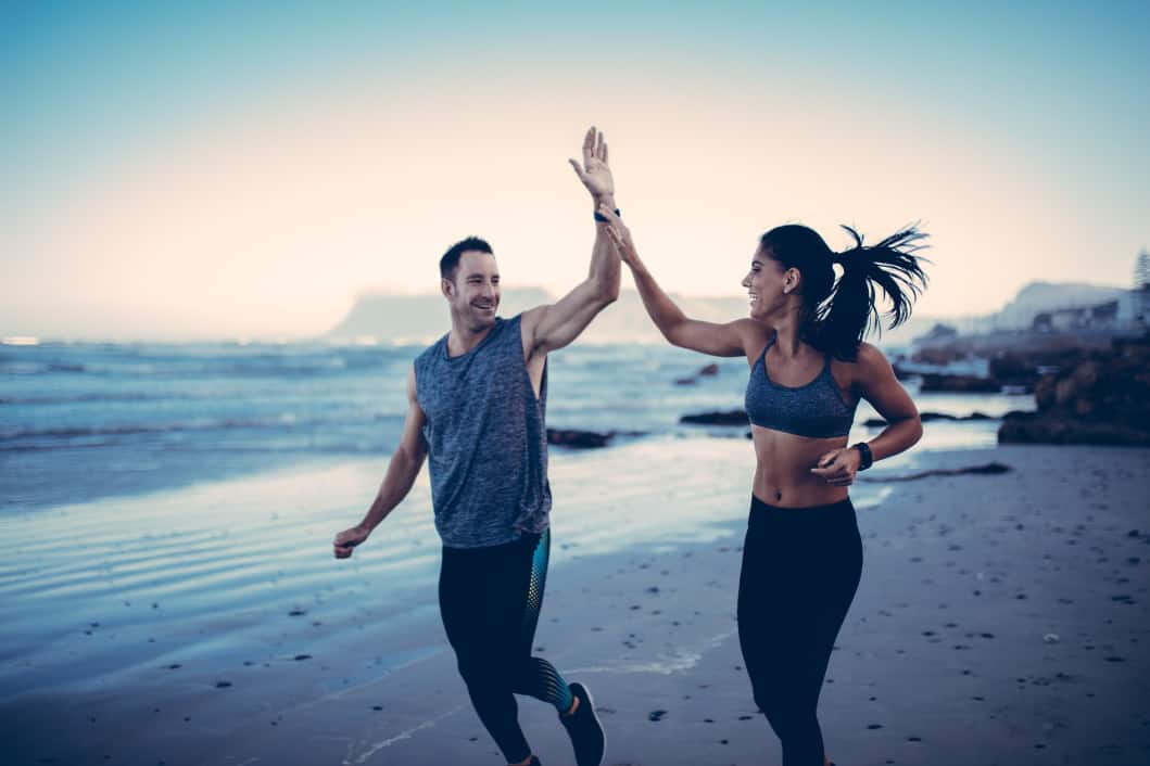 A man and woman jogging on a beach, smiling and giving each other a high five. They are wearing athletic clothing and the ocean waves are in the background, with a distant mountain visible under a clear sky.