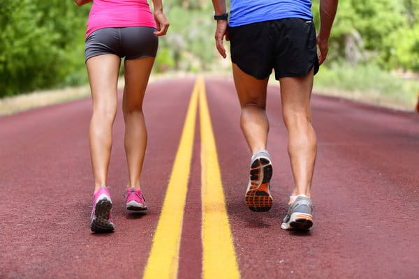 Two people walking on a paved road with a yellow centerline. The person on the left wears a pink shirt, dark shorts, and pink sneakers, while the person on the right wears a blue shirt, black shorts, and gray sneakers. The road is surrounded by greenery.