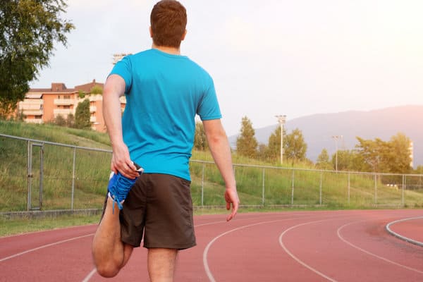 A man wearing a blue shirt and black shorts stretches his left leg by holding his ankle while standing on a red track. He is facing away from the camera with a grassy area and buildings in the background. The sky is bright and hazy.