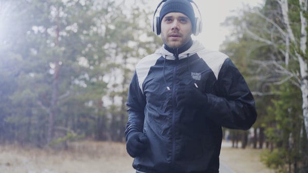 A man jogs outdoors on a clear day, wearing a black and white jacket, black gloves, a dark beanie, and large white headphones. He appears focused and determined, running along a path with trees in the background.