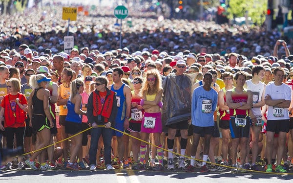 A large crowd of runners gathers at the starting line of a race. People of various ages and genders are wearing race bibs and athletic gear in bright colors. The atmosphere is energetic, with spectators and participants eagerly awaiting the start.