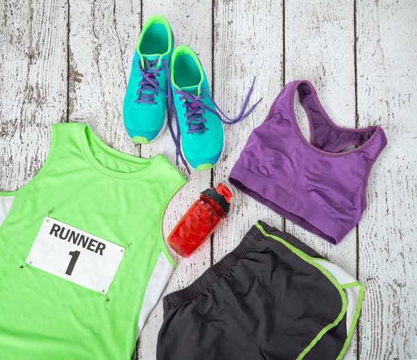 Top view of running gear on a wooden surface, featuring a pair of bright blue running shoes, a green tank top with a "Runner 1" bib, a purple sports bra, black shorts with green trim, and a red water bottle.