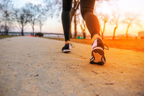 Close-up of a person walking on a trail during sunset. The focus is on their legs and shoes, with the background showing leafless trees and a scenic view. The lighting suggests a serene, calm moment in an outdoor setting.