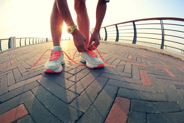 A person is bending down to tie the neon orange laces of their running shoes on a paved pathway with a sunrise in the background. The person is wearing a yellow wristwatch, and a railing is visible along both sides of the path.