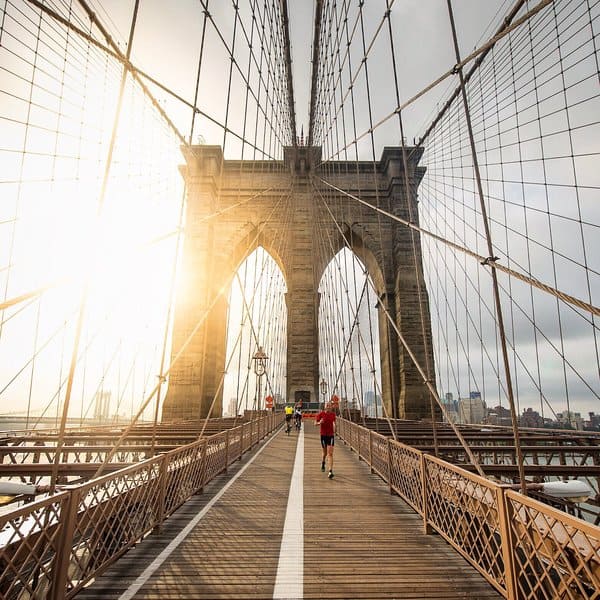 A person in athletic wear jogs across the Brooklyn Bridge during sunrise. The bridge's iconic cables frame the stone arches, and the early morning light creates a warm, glowing effect. The city skyline is faintly visible in the background.