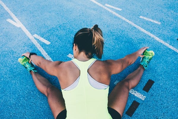 A person with a ponytail and wearing a bright yellow tank top and black shorts is seated on a blue track, stretching their legs apart and reaching to touch their green sneakers with both hands. White lines of the track are visible in the background.