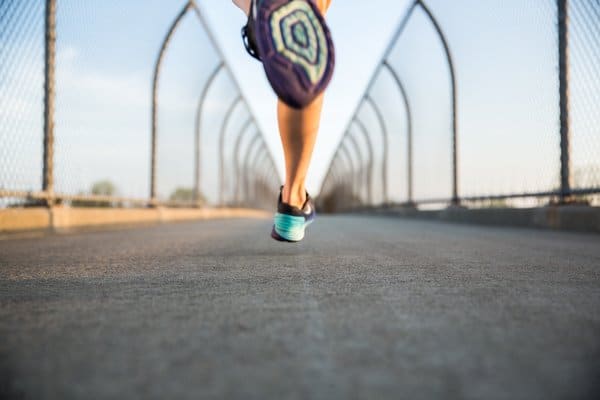 Close-up view of a person's legs in running shoes, captured mid-stride on a bridge with wire mesh fencing on both sides. The background displays a bright blue sky, giving the scene a sense of motion and freedom.