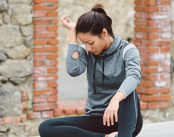 A woman in athletic wear sits outdoors in a thoughtful pose, with her elbow resting on her knee and her other hand touching her head. She is dressed in a gray long-sleeve top and black leggings, and she appears to be resting after exercise. The background features stone and brickwork.