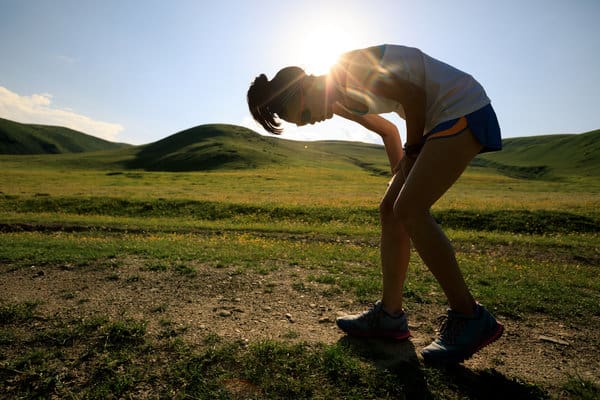 A person in athletic wear bends forward, hands on knees, seemingly fatigued while jogging along a dirt path in a grassy, sunlit landscape with gentle hills in the background. The sun shines brightly, creating a slight lens flare effect.