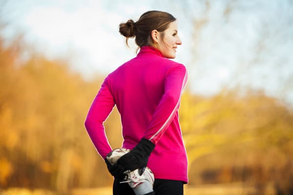 A person with their hair in a bun, wearing a bright pink long-sleeved top and black gloves, stands outdoors in a park during fall. They are smiling and stretching by holding one leg behind them. The background features blurred autumn foliage.