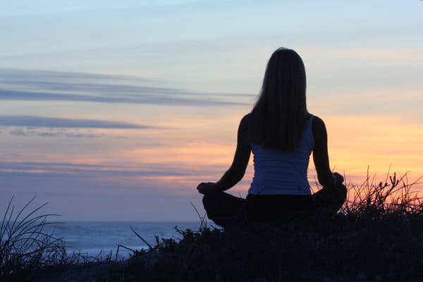 A person sits cross-legged facing the ocean, meditating at sunset. Tall grass surrounds the figure, and the sky is painted with soft hues of blue, orange, and pink as the sun sets in the background. The scene evokes calm and serenity.