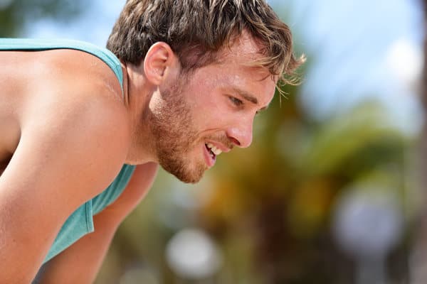 A man with light brown hair and a light beard, wearing a teal tank top, appears to be catching his breath and leaning forward. The background is a clear sunny day with blurred greenery and sky.