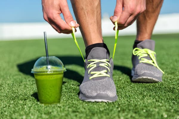 A person is tying neon green shoelaces on gray athletic shoes while kneeling on a grass field. A green smoothie in a plastic cup with a black straw is placed to the side. The day appears sunny with a clear blue sky in the background.