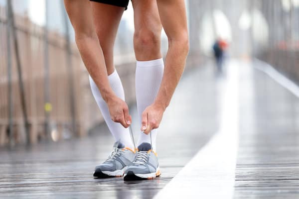 A person in athletic attire bends down to adjust their gray running shoes on a wet, wooden bridge. They are wearing knee-high white compression socks and black shorts. The background is blurred, but cage-like fencing and another distant figure are visible.