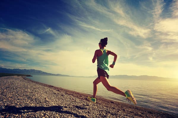 A person wearing a tank top, shorts, and running shoes is jogging along a pebbled path beside a calm lake. The sky above is partly cloudy with the sun casting a warm glow, creating a serene and picturesque setting.