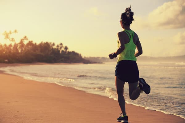 A person in a neon green tank top and black shorts is jogging along a sandy beach at sunset. The scene features gentle waves, a distant tree line, and a sky with scattered clouds tinted by the setting sun.