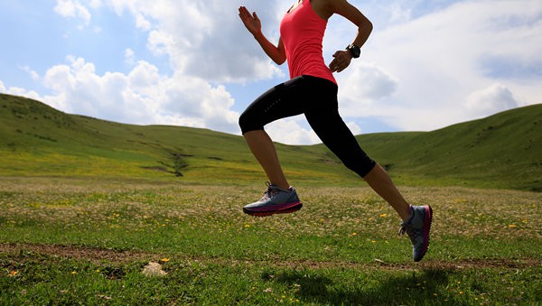 A person in a bright pink tank top, black leggings, and gray running shoes is mid-stride while running through a grassy, flower-filled field with rolling hills in the background under a partly cloudy sky.