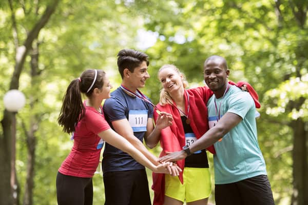 Four people, wearing athletic gear and participant numbers, stand in a circle with their hands stacked together in a park. They are smiling and appear to be celebrating after a run or athletic event. Trees and greenery are visible in the background.