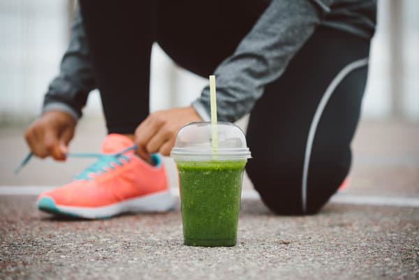 A person wearing a gray long-sleeve top and black leggings is tying the laces of bright orange running shoes. In the foreground, there is a green smoothie in a plastic cup with a straw, placed on the ground.