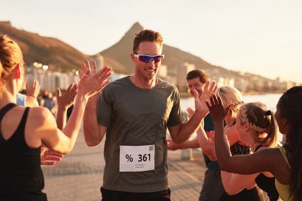 A runner wearing a green shirt and race number 361, sporting sunglasses, high-fives supporters gathered along the seaside race route. The sun is setting, casting a warm glow, with a mountainous backdrop and cityscape in the distance.