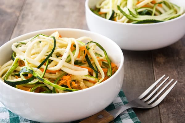 Two white bowls filled with zoodle salad, containing spiralized zucchini, carrots, and other vegetables, are placed on a wooden table. A fork rests on a green and white checkered cloth next to one of the bowls.