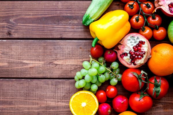 A colorful assortment of fruits and vegetables is arranged on a wooden surface. Items include grape tomatoes, yellow bell pepper, pomegranate halves, green grapes, orange halves, zucchinis, and cherries. The left part of the image shows the wooden surface.