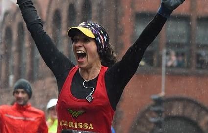 A person enthusiastically celebrating with arms raised, wearing a red tank top with "Carissa" written on it and a Wonder Woman logo, a black long sleeve underneath, and a colorful visor. It's raining, and blurred figures and buildings are visible in the background.