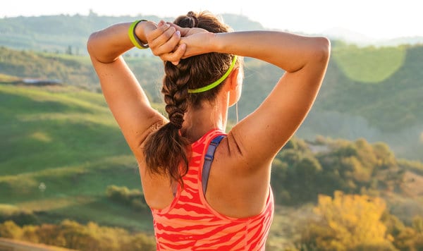 A woman with a braid, wearing a neon headband and a pink athletic sleeveless shirt, stands outdoors with her arms raised, adjusting her hair. She overlooks a scenic, sunlit landscape of rolling green hills and trees.