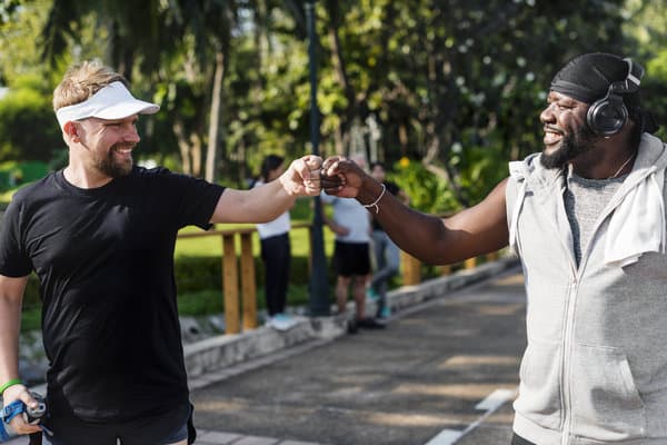 Two men are smiling and giving each other a fist bump outdoors. One wears a white visor and black athletic gear, and the other wears a black beanie, gray sleeveless hoodie, and headphones. They are standing on a paved path with trees and people in the background.