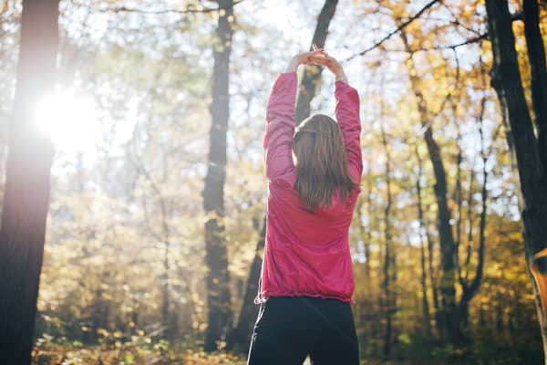 A person with long hair, wearing a pink jacket and dark pants, stretches with arms overhead in a sunlit forest with tall trees and autumn foliage. The scene captures a calm, nature-filled moment on a sunny day.