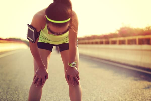 A person in athletic wear, including a headband and armband, is bent over with hands on knees, appearing fatigued while standing on an empty road under a bright sky.