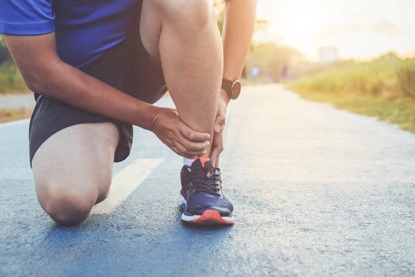 A person wearing a blue shirt and black shorts kneels on one knee to tie their running shoe on a paved path. The scene is outdoors with grass on the sides of the path and sunlight in the background. The person is also wearing a smartwatch.