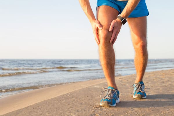 A person in blue athletic shorts and blue sneakers is standing on the beach, holding their knee in apparent discomfort. The background features a calm ocean and a clear sky. The person is wearing a smartwatch on their left wrist.