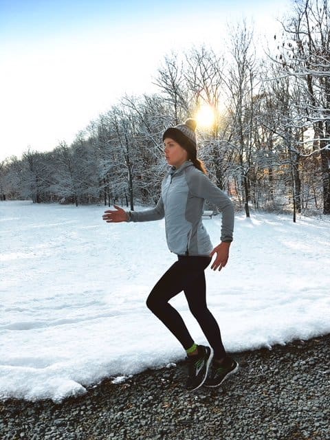 A person dressed in winter running gear, including a hat and gloves, jogs along a snow-covered path surrounded by bare trees. The sun is shining through the trees, creating a serene winter atmosphere.