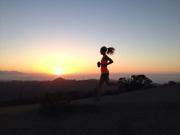 A silhouette of a person jogging on a trail at dusk. The sun is setting behind mountains in the background, casting an orange glow across the sky. The runner's hair is tied back in a ponytail, and they wear a tank top and shorts.