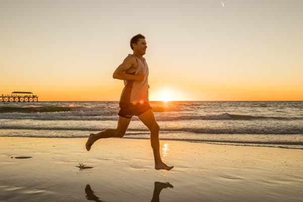 A man runs barefoot along a beach at sunset, with the sun low on the horizon casting a warm glow. He is wearing a tank top and shorts, and the ocean waves gently lap at the sandy shore beside him. A pier extends into the water in the background.