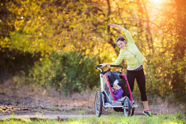 A woman stretches her arm overhead while holding onto a stroller with a child inside. The setting is outdoors in a park with autumnal trees and a warm, golden glow from the sun shining through the foliage. The child looks up at the woman, who is dressed in athletic wear.